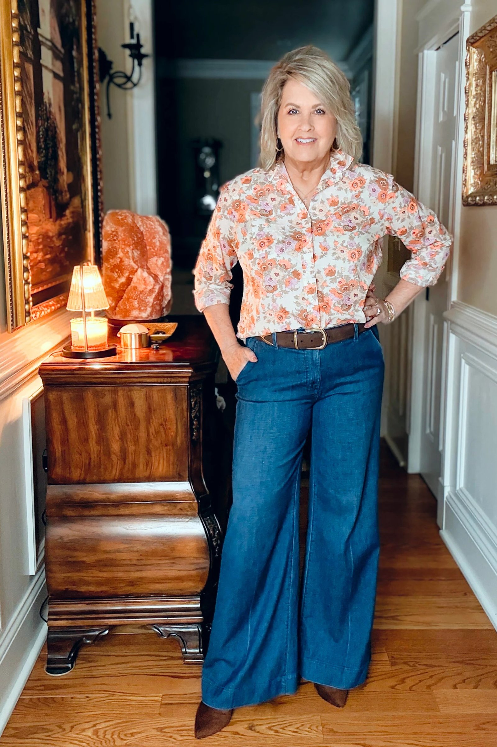 Woman over 50 wearing Floral print blouse tucked into dark wide-leg jeans with a brown belt, standing in a home hallway.