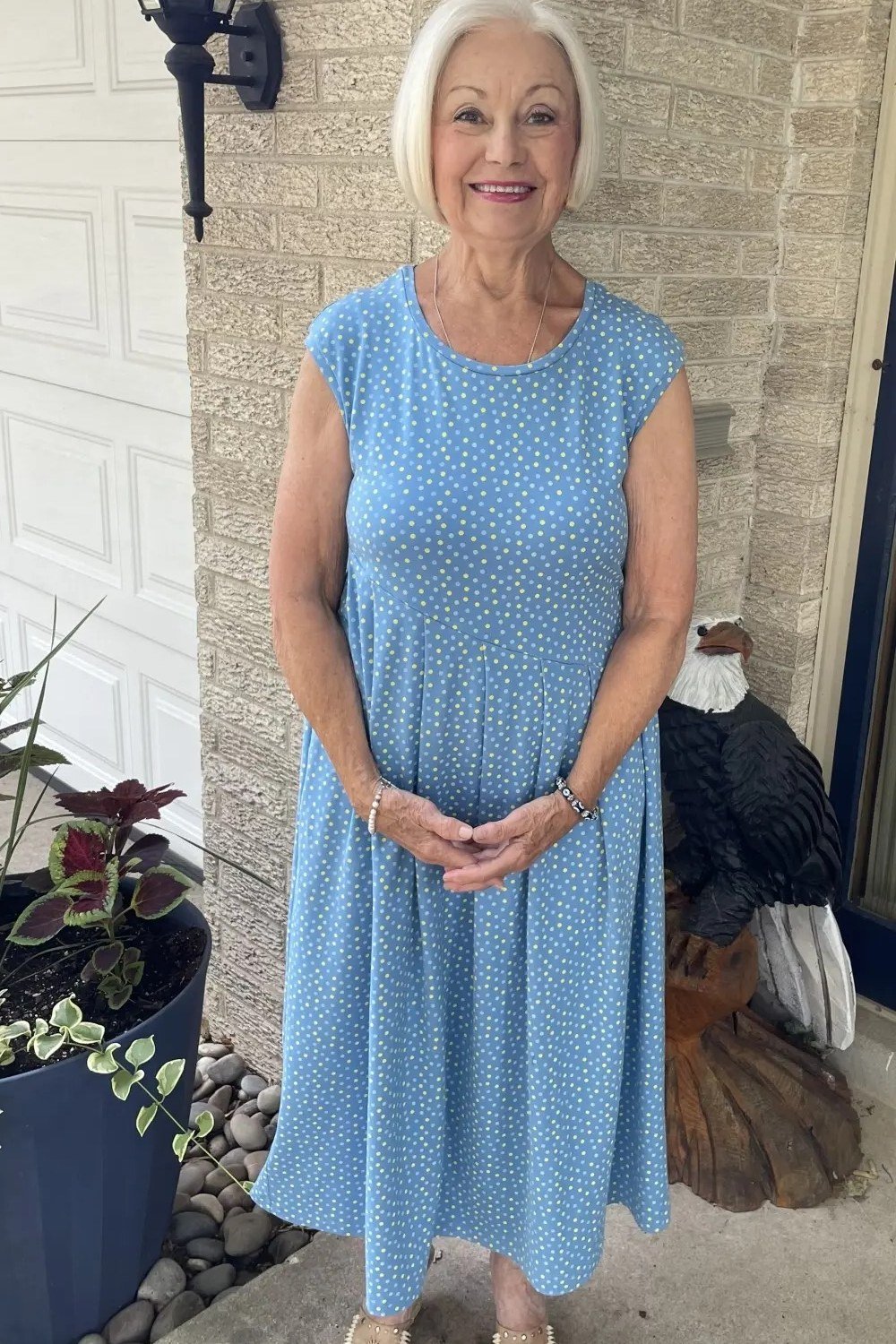 Older woman with white hair in a sleeveless light-blue knee-length dress, standing in front of a house, front view - summer dress for women over 50