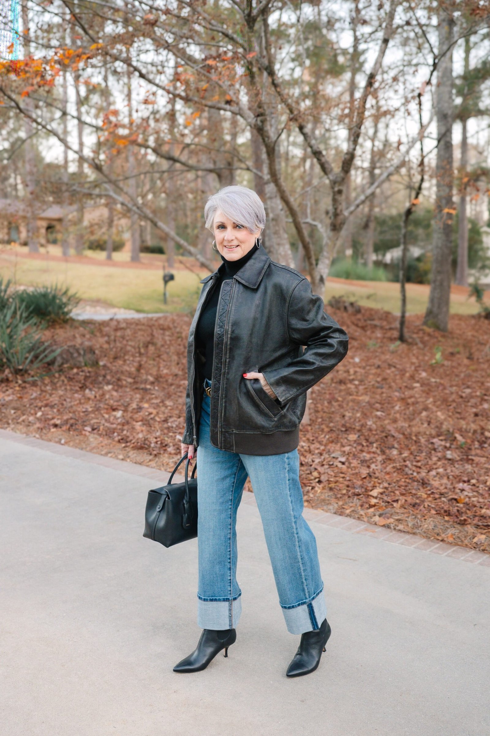 Woman over 60 wearing black leather jacket, light-wash jeans, black handbag, autumn park
