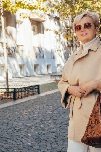 older woman in beige trench coat with cream top and white wide pants