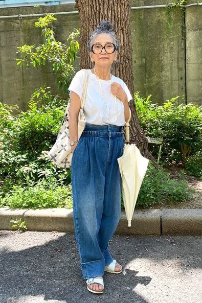 Woman over 50 wearing White tee with wide-leg blue jeans, holding a folded umbrella on a city street.