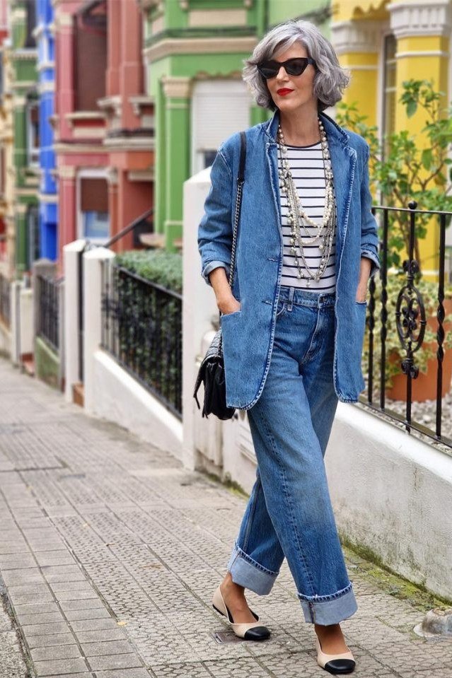 Woman over 50 wearing Chambray shirt over a striped tee with cuffed jeans on a colorful street.