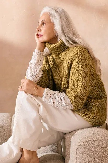Mustard cable-knit turtleneck sweater with cream trousers seated on sofa, cozy sweater outfit idea for women over 70