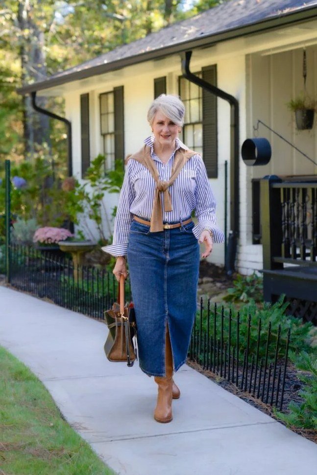 Woman over 50 wearing Striped shirt with a denim maxi skirt and brown boots on a garden path.