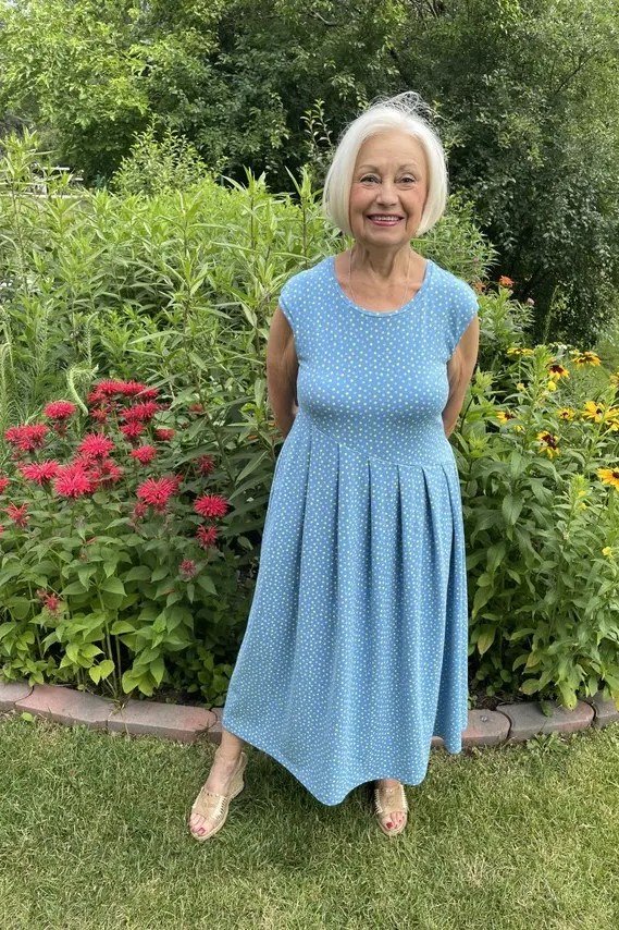 Older woman in a light-blue sleeveless knit midi dress, standing in a flower garden - summer dress for women over 50