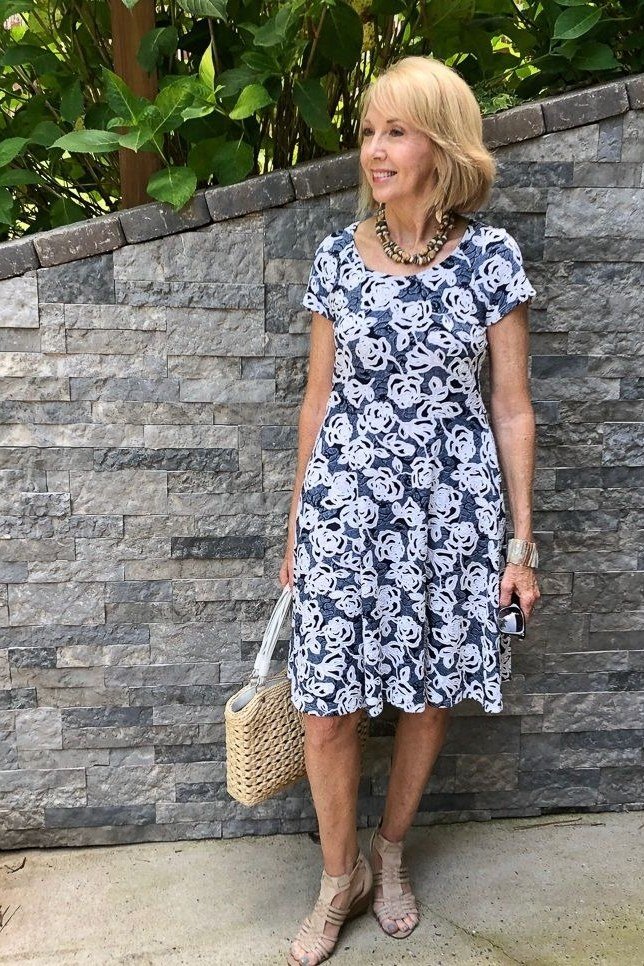 Blonde woman in a navy-blue floral short-sleeve midi dress with a straw bag, stone wall - summer dress for women over 50