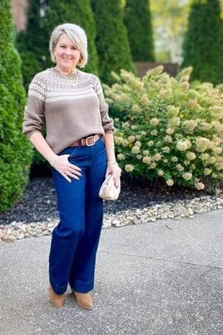 Woman over 60 wearing taupe sweater, medium blue jeans, bushes background