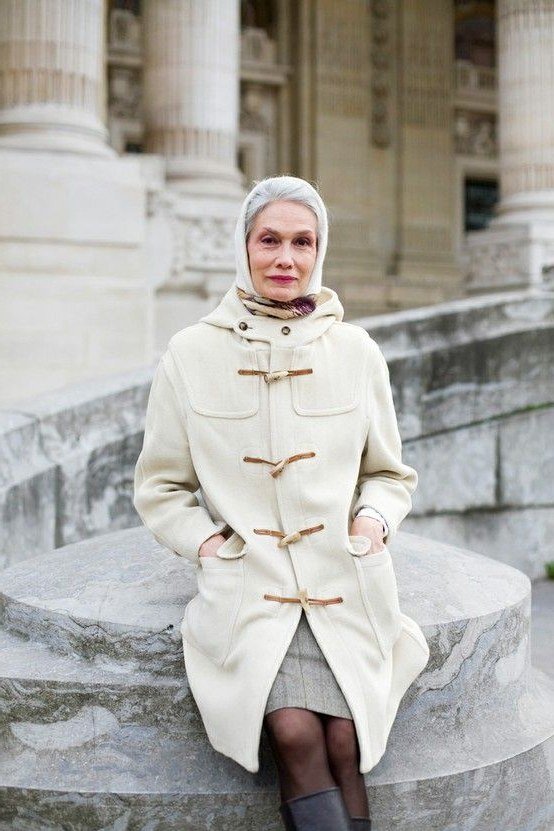 Stylish winter outfit for women over 60: cream duffle coat with gray scarf seated on stone steps