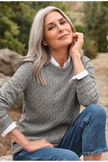 Grey cable-knit crew sweater with jeans seated on rocks by lake, cozy sweater outfit idea for women over 70