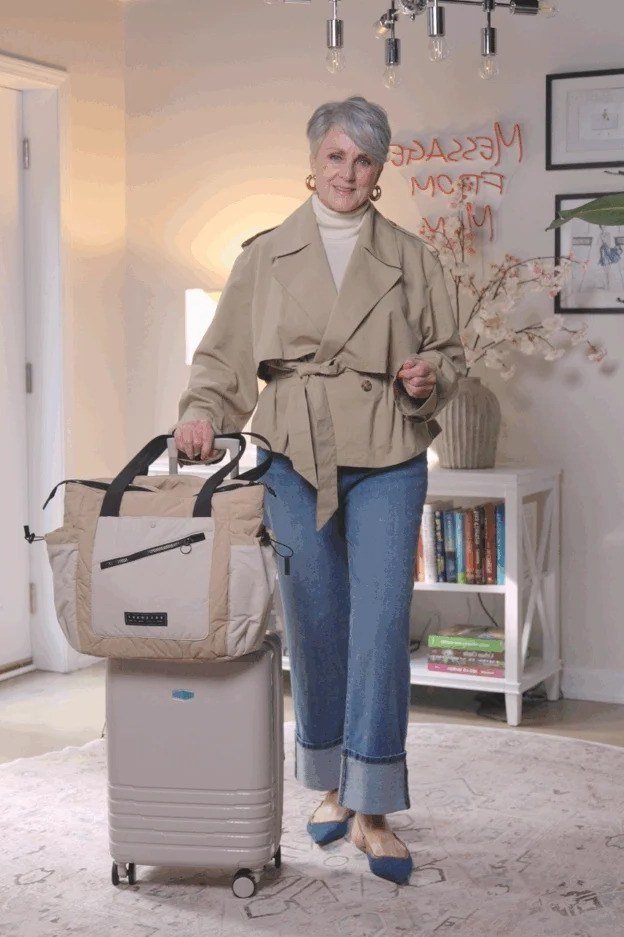 Woman over 50 in beige belted travel jacket with jeans and suitcase