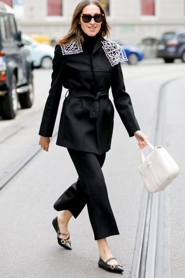 Black belted coat black trousers white bag, woman on city street - business casual work outfit for women over 50