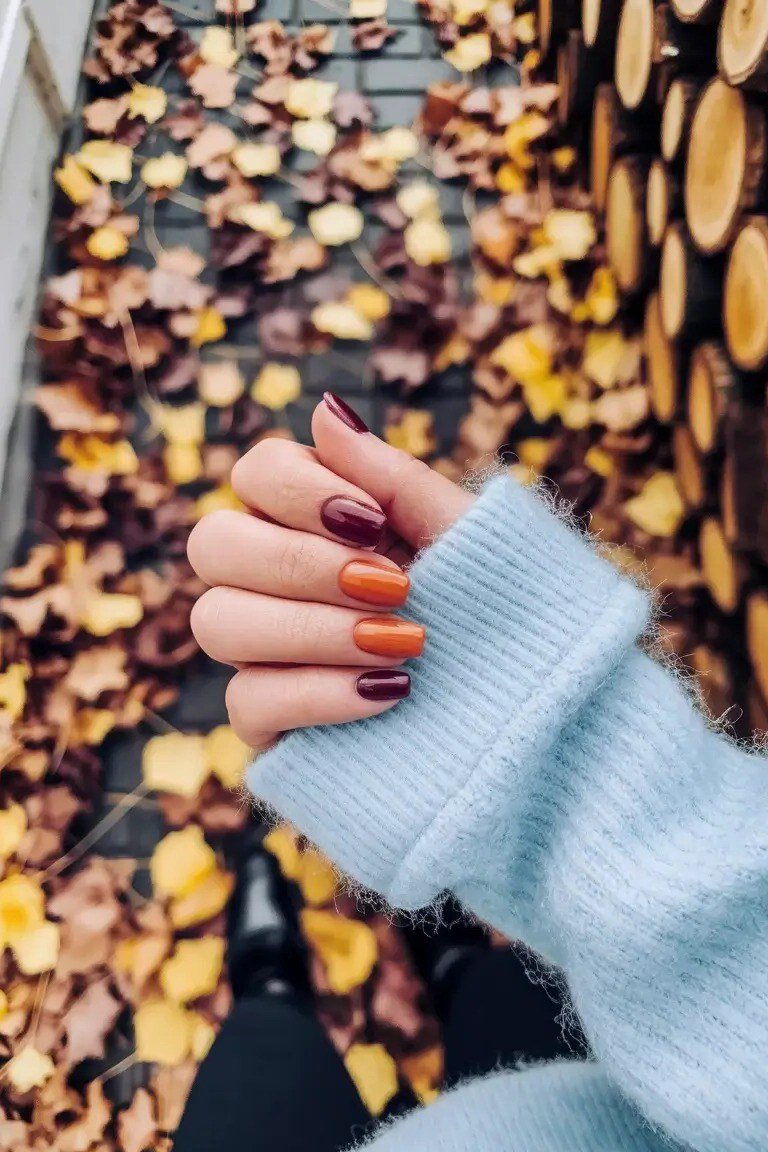 Multi-color fall nails in red, mustard and orange held above a bed of scattered autumn leaves.