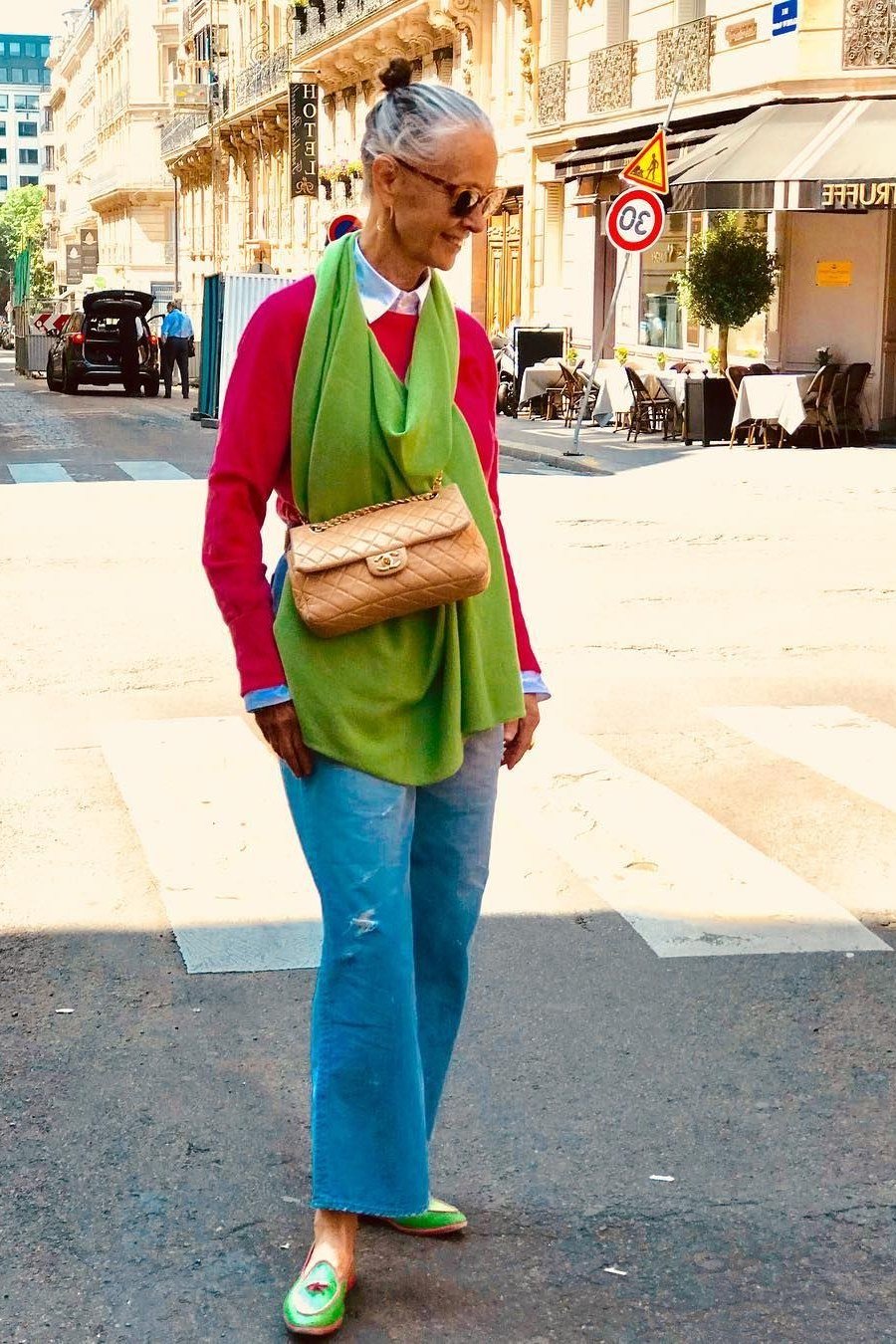 Woman over 50 wearing Pink sweater with a green scarf, blue jeans and green flats on a city street.