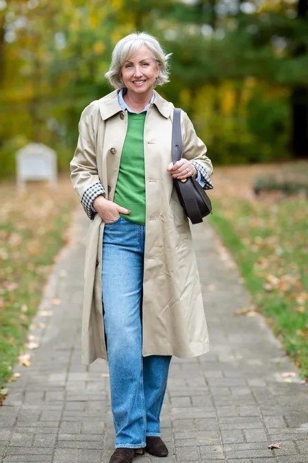 Classic beige trench coat over green sweater with blue jeans, woman over 50