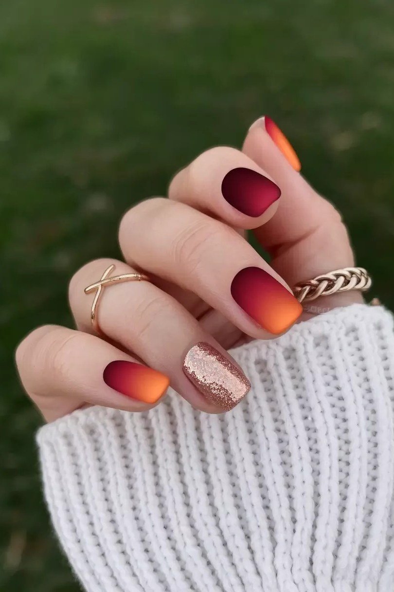 Sunset ombre nails blending orange to red with a gold glitter accent nail, glossy and vibrant.