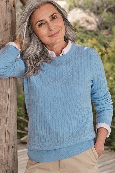 Sky-blue cable-knit sweater with tan trousers by wooden wall, cozy sweater outfit idea for women over 70