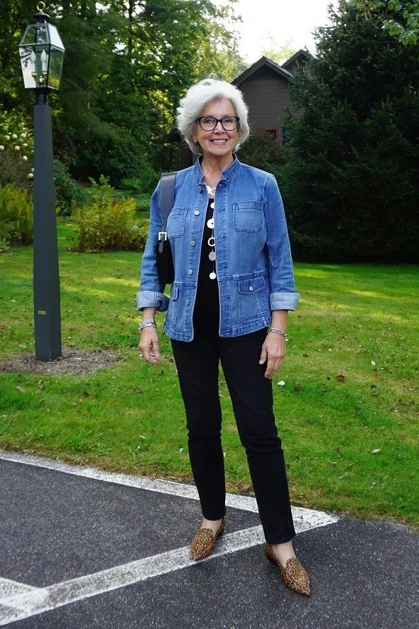 Woman over 50 wearing Blue denim jacket with black pants outdoors in a park.