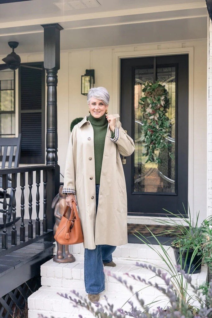 Woman over 50 in beige trench coat with forest green turtleneck on porch