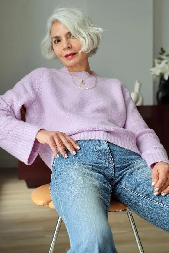 Woman over 60 wearing lavender sweater, light jeans, seated indoors