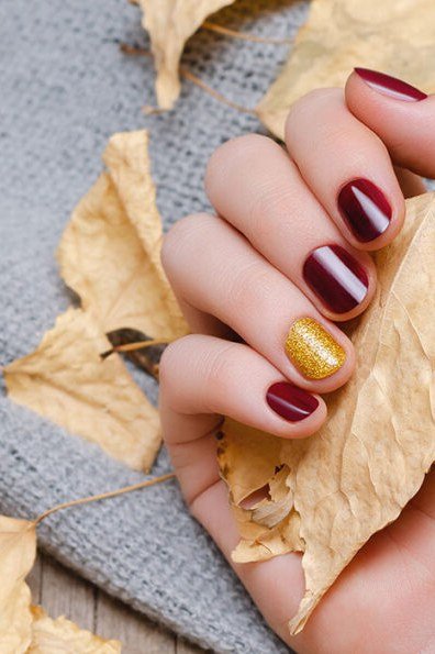 Burgundy oxblood solid with gold leaf accent on one nail, short oval, glossy, hand holding dried autumn leaves, fall nail color for women over 40