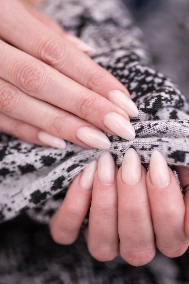 Pale neutral pink almond nails with a glossy finish, resting in a patterned knit sweater.