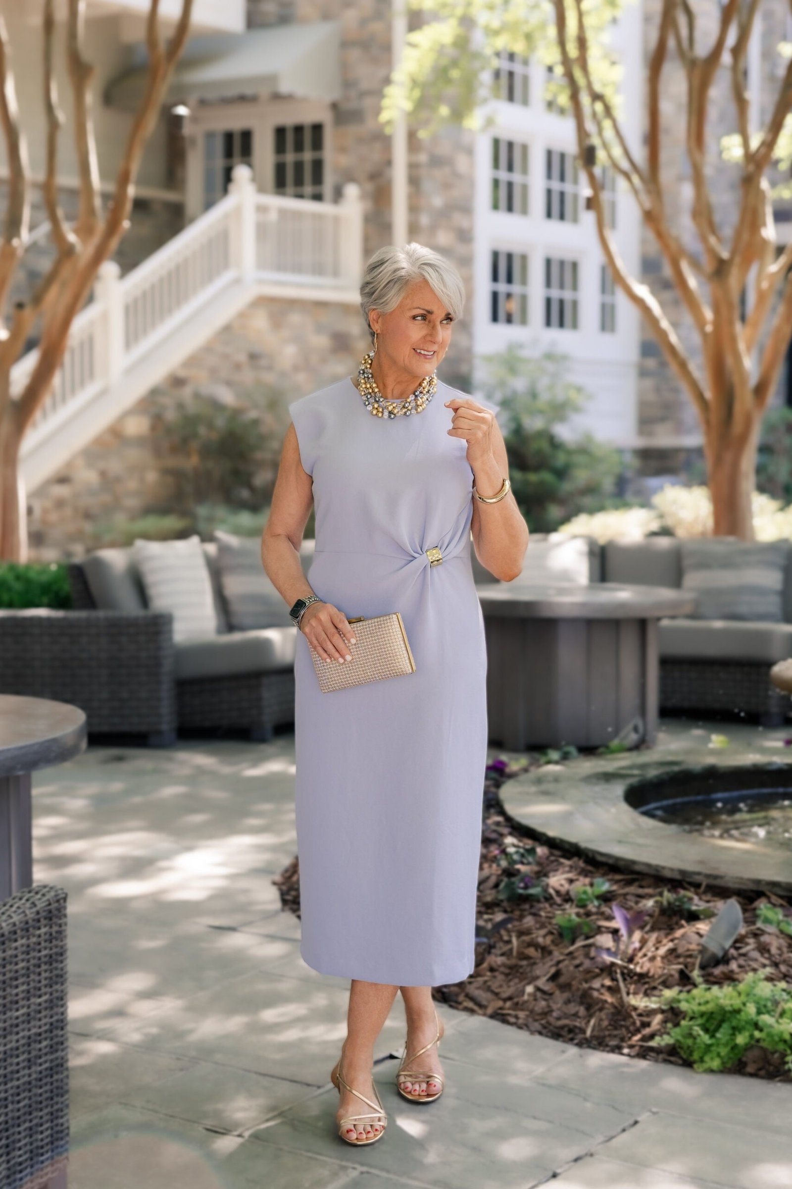 Older woman in a lavender cap-sleeve midi dress with a straw clutch, courtyard - summer dress for women over 50