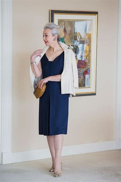 Gray-haired woman in navy sheath dress with white cropped jacket and clutch