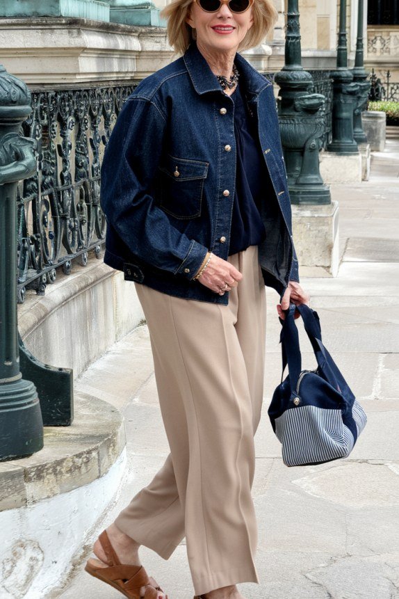 Woman over 50 wearing Dark denim jacket with navy top, beige wide pants and blue tote in the city.
