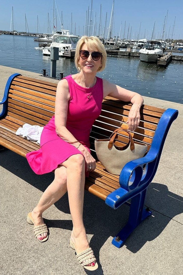 Older woman in a sleeveless fuchsia fit-and-flare dress, sitting on a park bench by the water - summer dress for women over 50