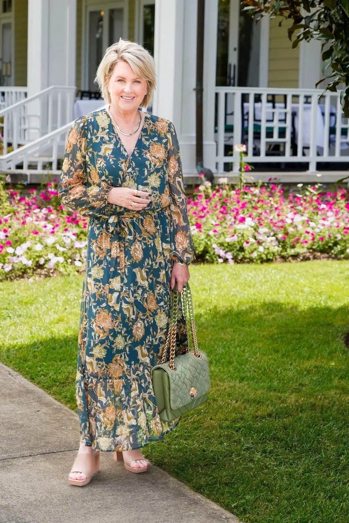 Older woman in a long blue floral maxi dress with long sleeves, holding a sage-green handbag, garden - summer dress for women over 50