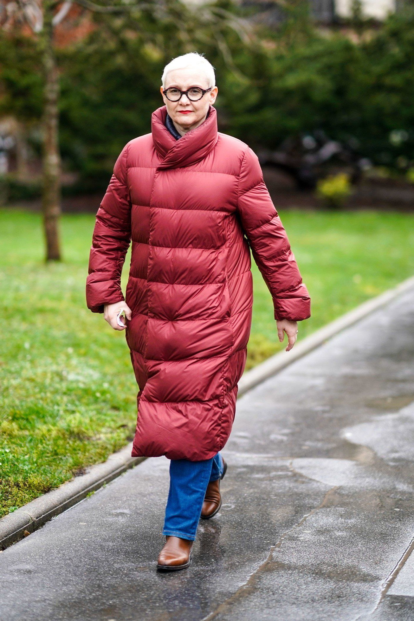 Stylish winter outfit for women over 60: long red puffer coat with jeans on a park path