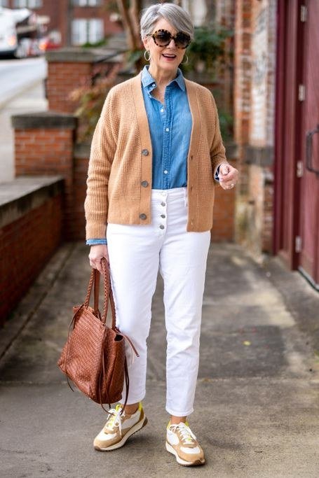 Woman over 50 wearing Tan cardigan over a blue shirt with white jeans and brown tote on the street.