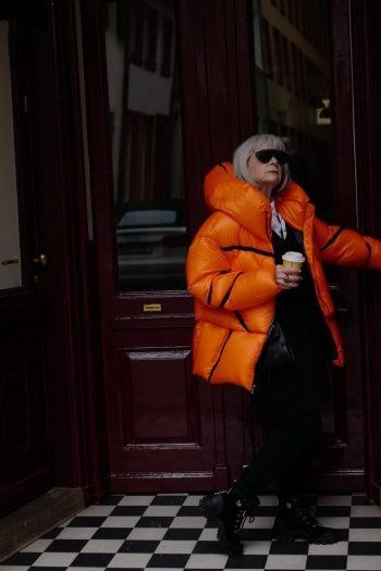 Stylish winter outfit for women over 60: orange puffer jacket with red scarf holding a coffee cup in a dark doorway