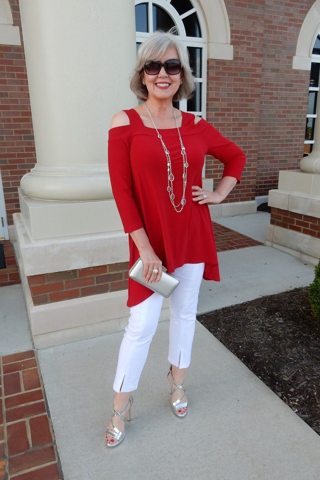 Red cold-shoulder tunic, white cropped pants, silver clutch and heels, brick exterior, chic night out outfit for women over 50