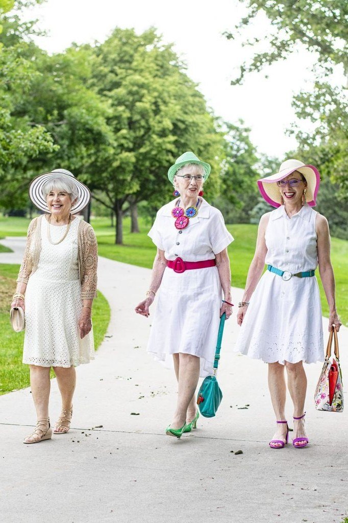 Three older women in long white dresses and hats strolling in a park - summer dress for women over 50