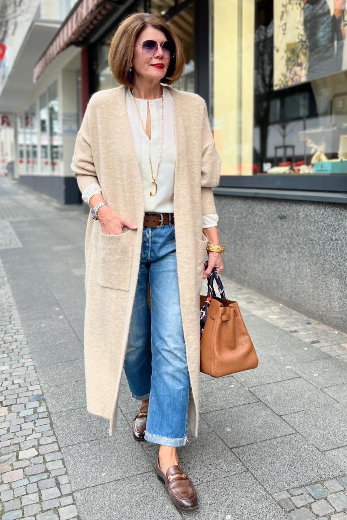 Long beige cardigan with cream top, blue jeans and cognac handbag