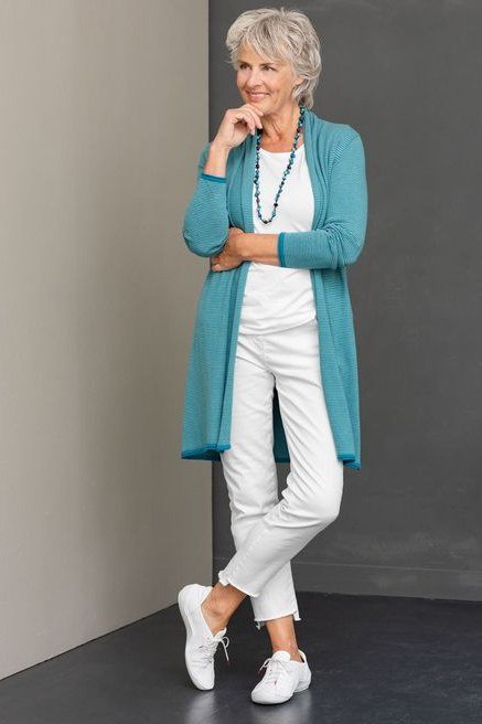 Woman over 50 wearing Turquoise cardigan with white top, white pants and white sneakers, studio shot.