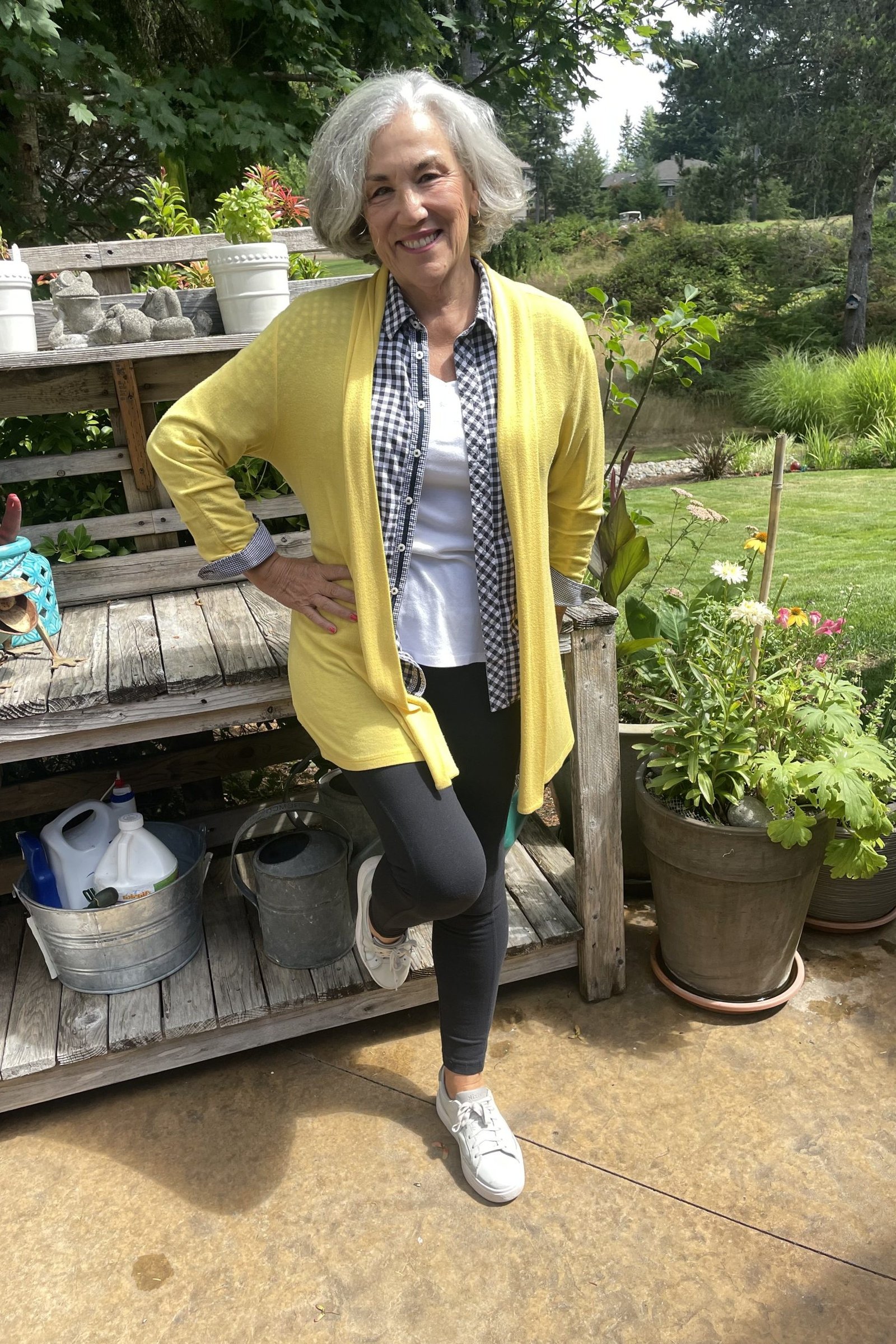 Woman over 50 wearing Yellow cardigan with checkered shirt, black pants and white sneakers in a garden.
