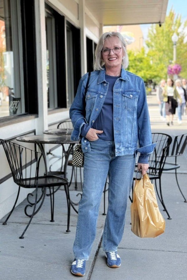 Woman over 60 wearing denim jacket, navy top, medium jeans, tan bag, cafe