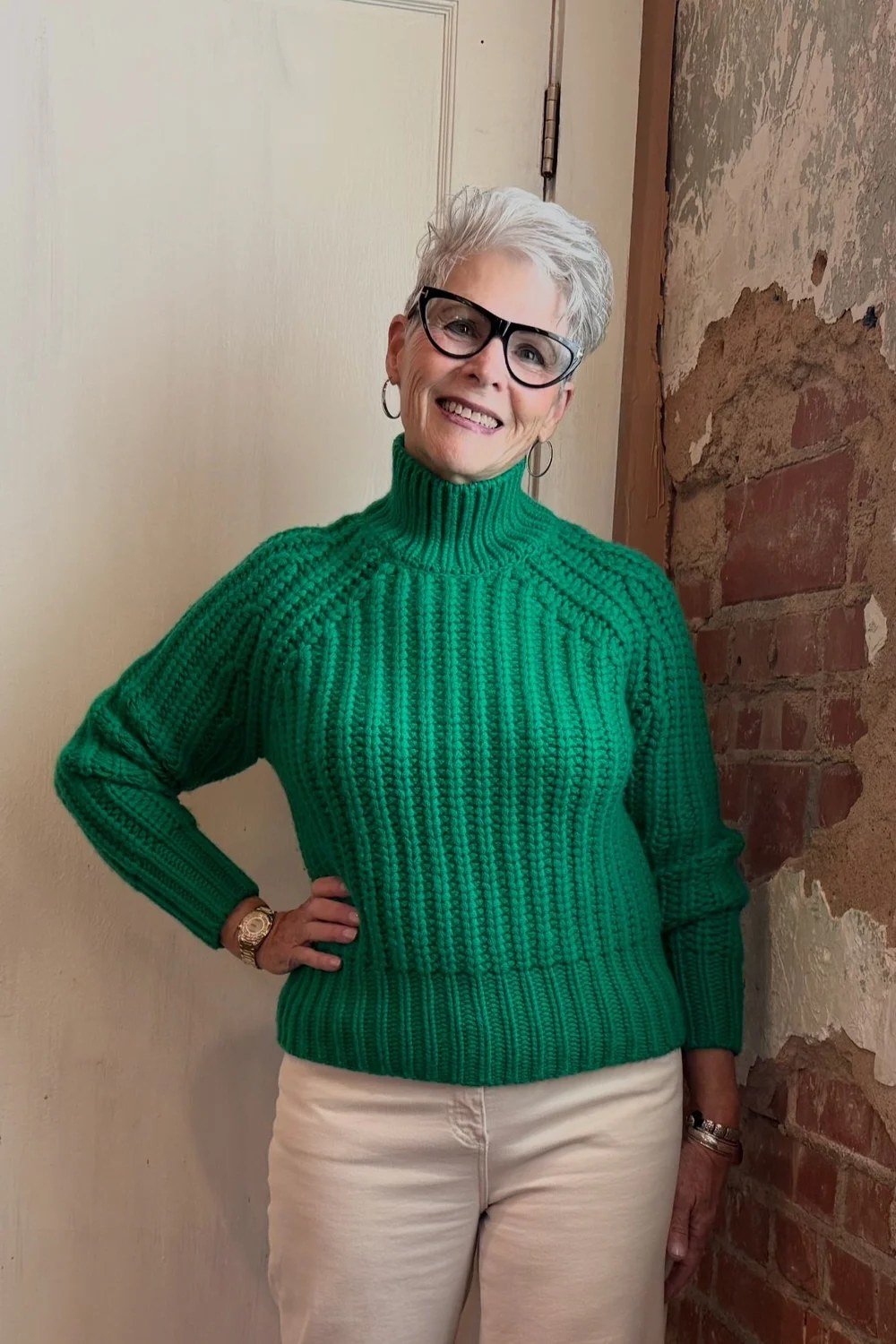 Stylish winter outfit for women over 60: green ribbed turtleneck sweater with cream trousers against a brick wall