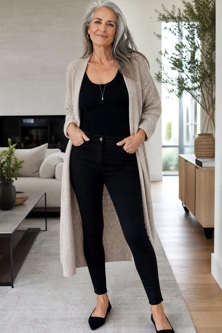 Woman over 50 wearing Long beige cardigan with black top and black jeans in a living room.