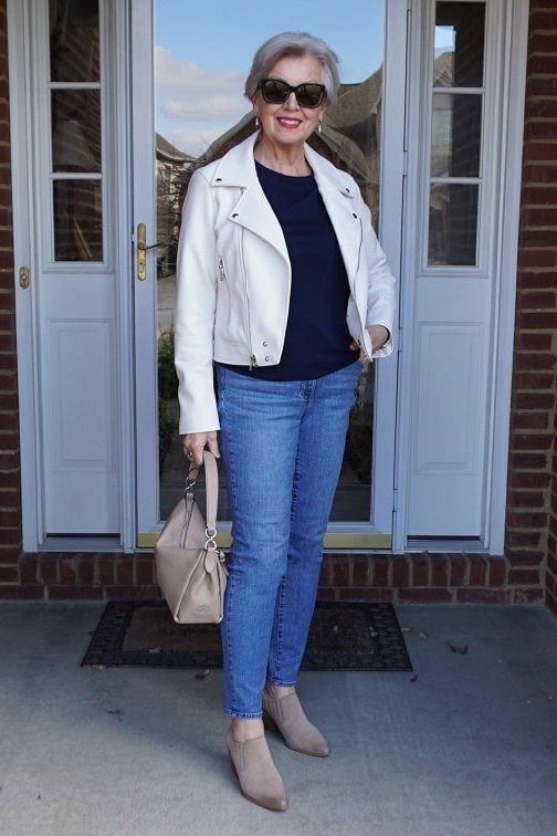 Woman over 60 wearing white moto jacket, navy top, medium jeans, brick doorway