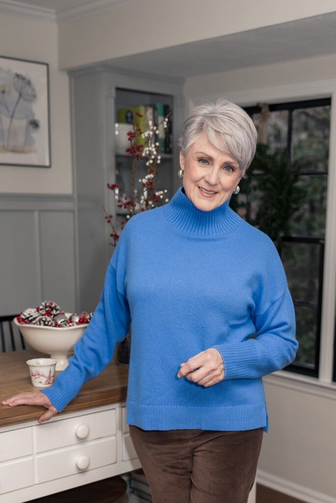 Bright-blue turtleneck sweater with brown trousers in kitchen, cozy sweater outfit idea for women over 70