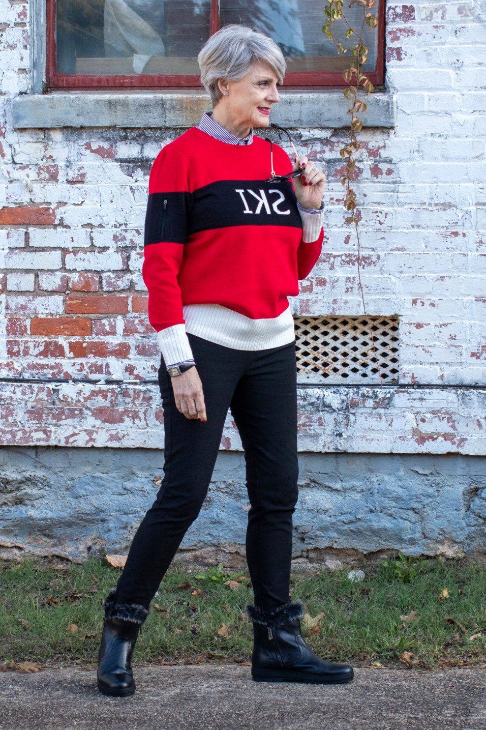 Stylish winter outfit for women over 60: red and black ski sweater with black jeans against a brick wall