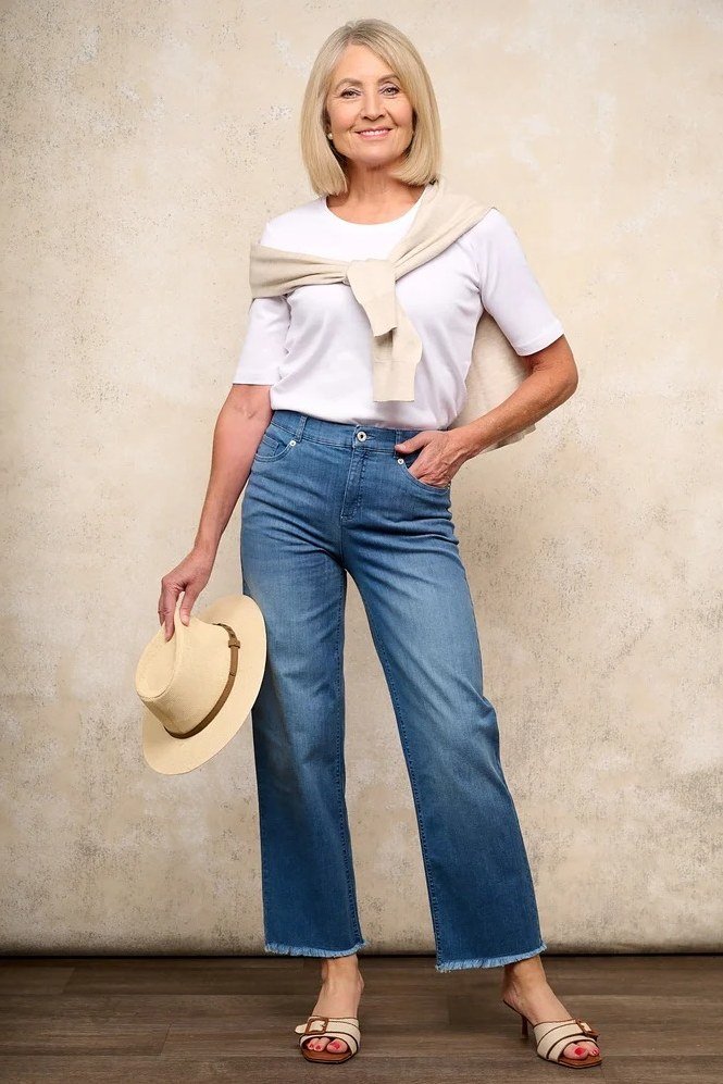Woman over 60 wearing white tee with sweater draped over shoulders, wide jeans, straw hat