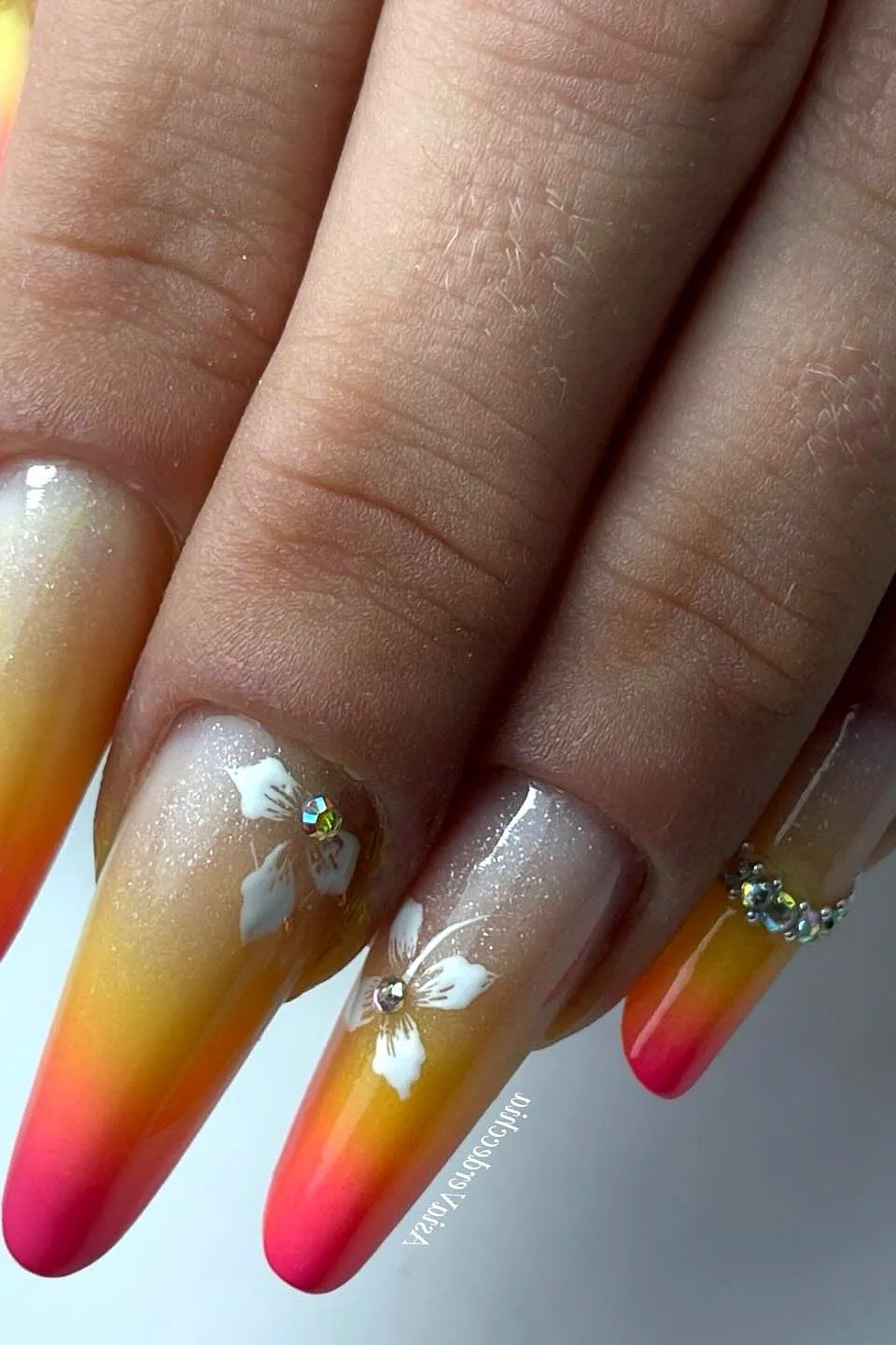 Sunset Ombre Stiletto Nails with Floral Art - Long stiletto nails glow with a yellow to hot pink sunset gradient, finished with white flower art, gold glitter, and tiny rhinestones near the cuticle. The glossy finish looks like tropical blooms at sunset.