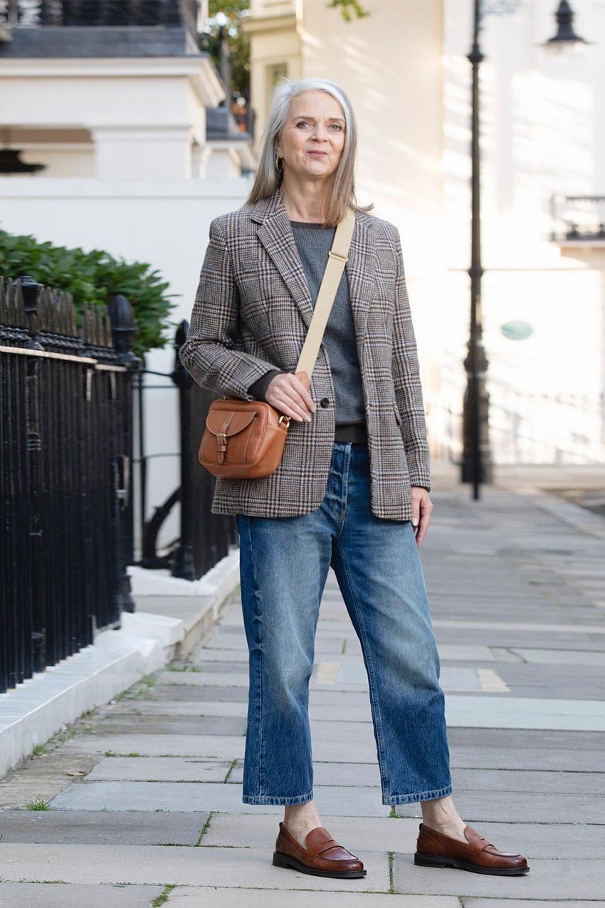 Woman over 50 wearing Houndstooth blazer with grey top, blue jeans, loafers and crossbody on a London street.