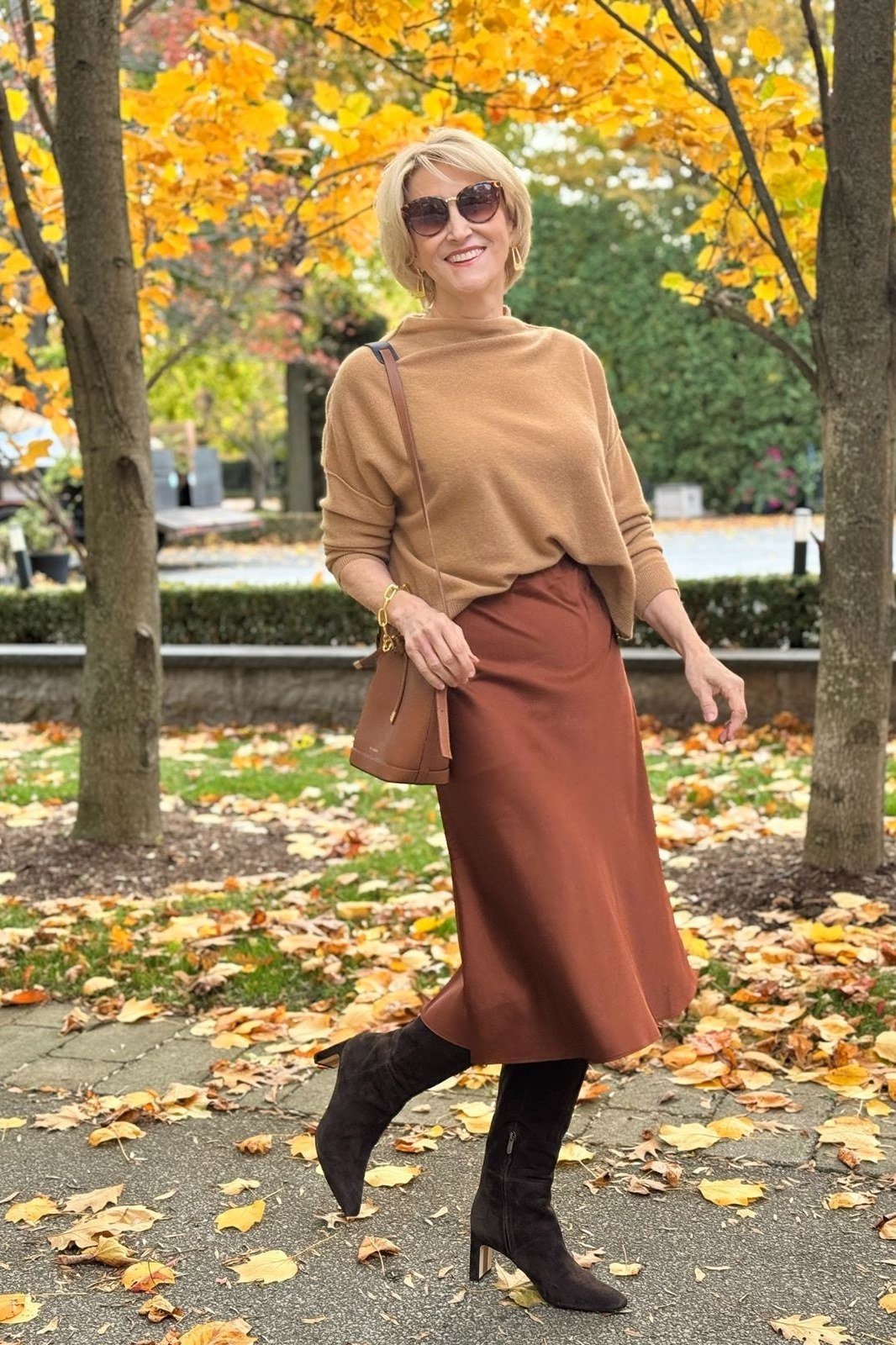 older woman in camel sweater with rust satin midi skirt and black boots, autumn leaves