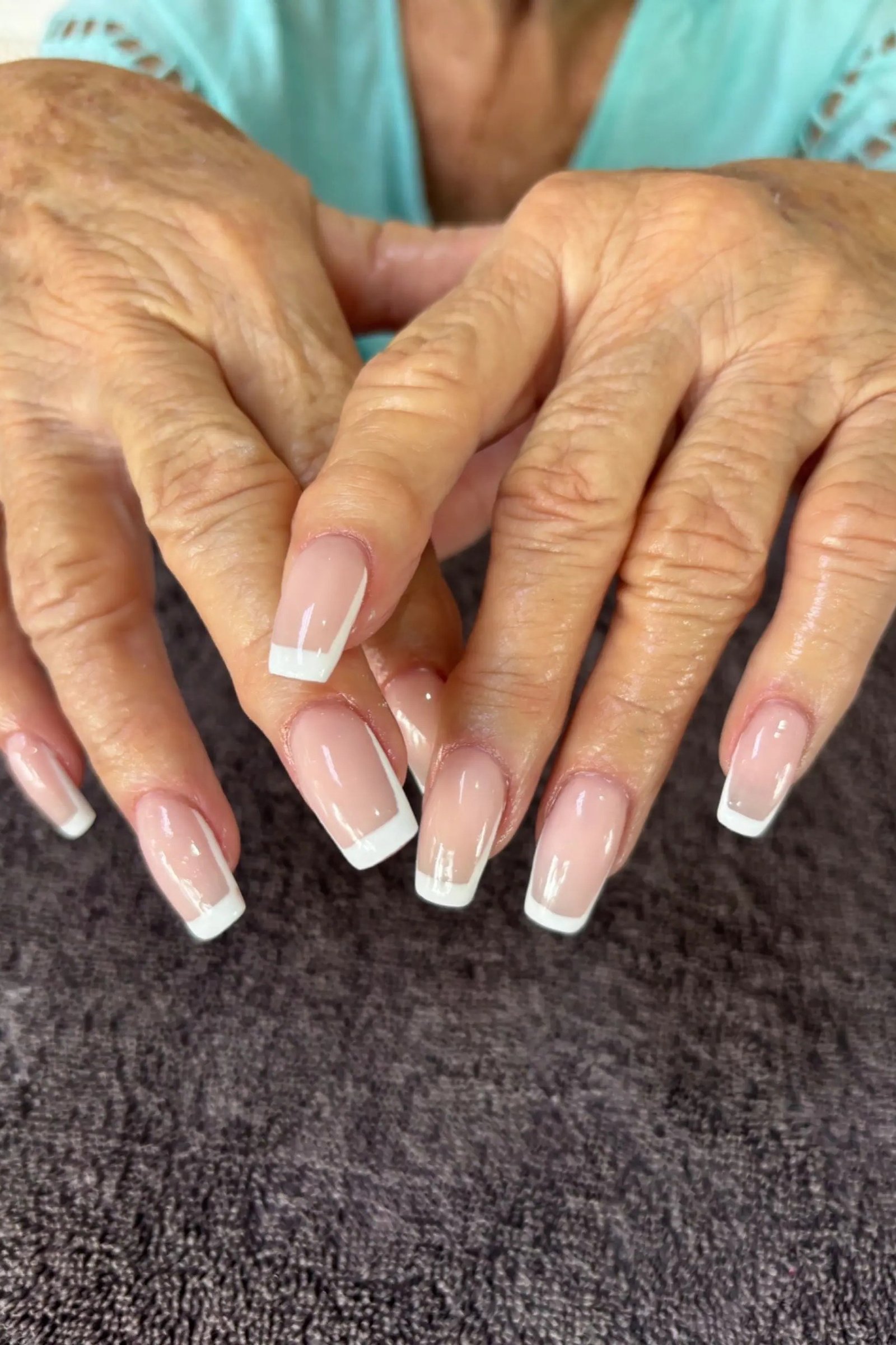 Nude pink french tip almond nails with a glossy finish on mature wrinkled hands.