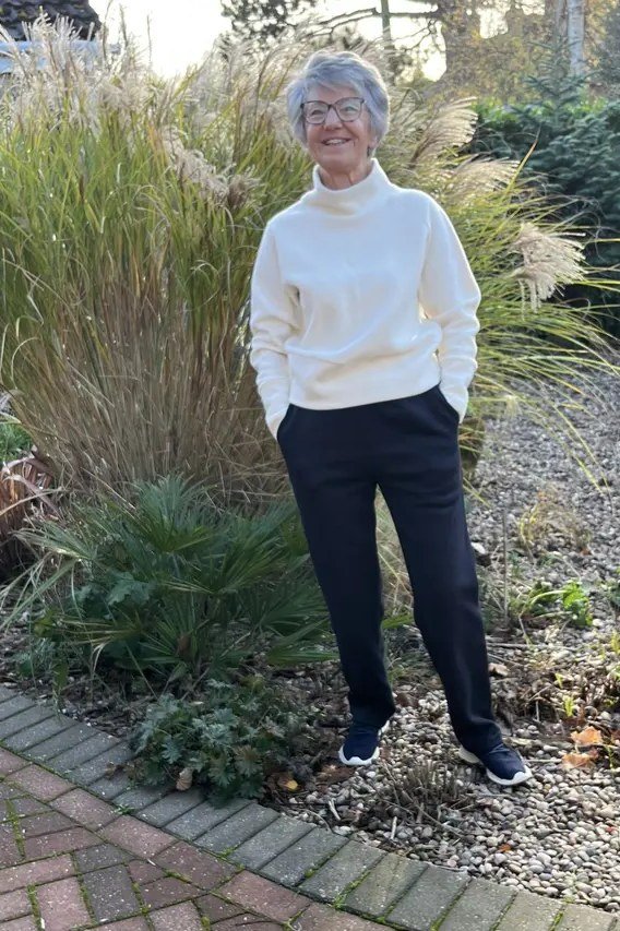 Stylish winter outfit for women over 60: white knit sweater with dark trousers on a garden path